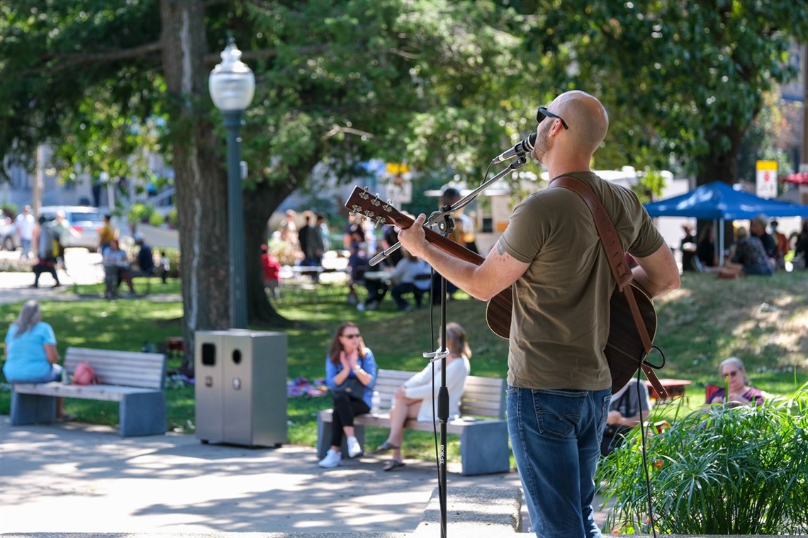 Person playing guitar on the Rotary Stage in Bronson Park at a Lunchtime Live Event. Food trucks, vendors, and event attendees in the background.