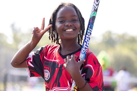 A photo of a child holding a hockey stick, smiling, and making a peace sign. 